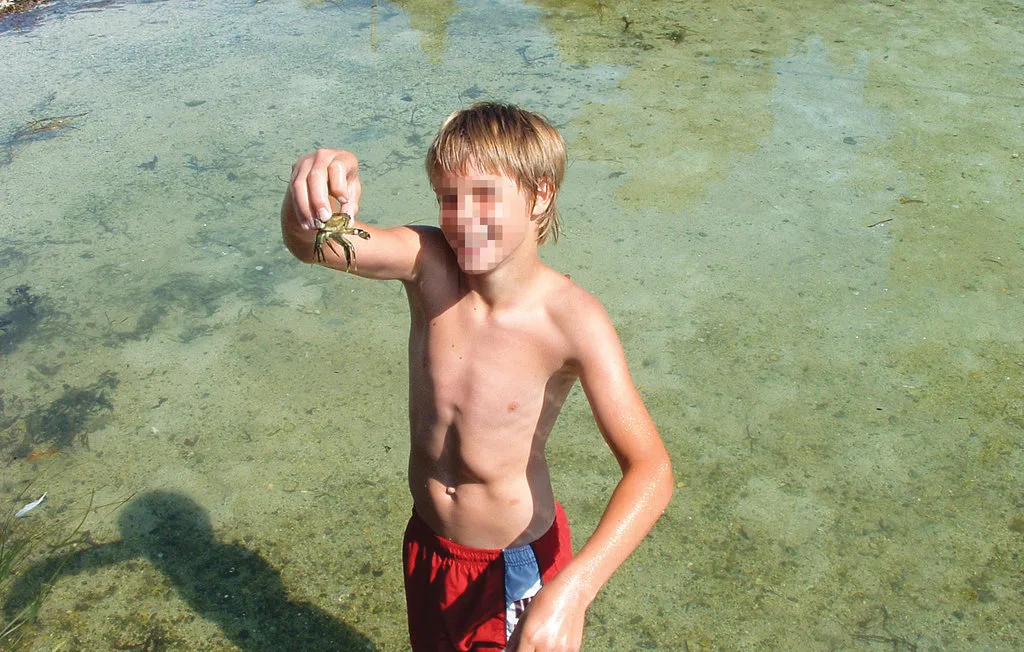 A child stands in shallow clear water, holding a small crab.