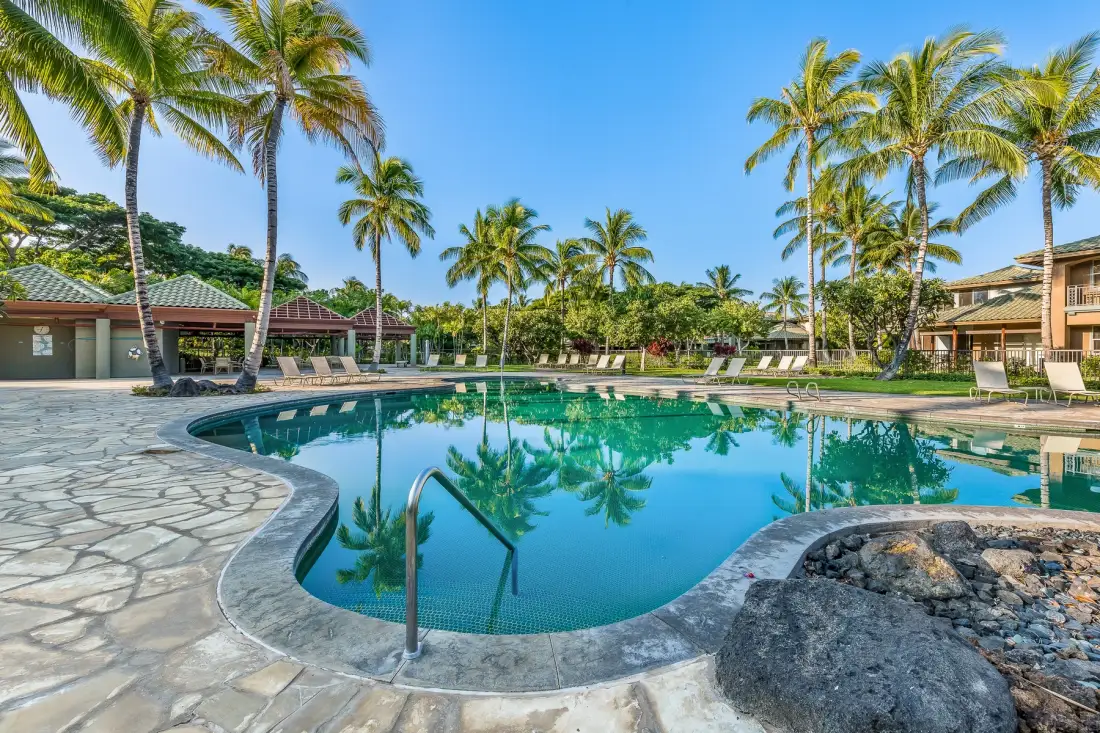 Una piscina de resort está rodeada de palmeras, con pavimento de piedra y asientos cercanos.