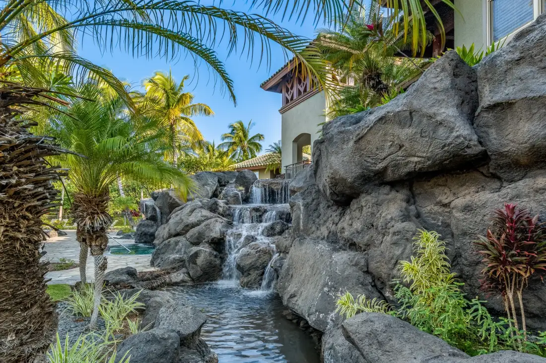 Un jardín con una cascada entre rocas que desemboca en una piscina bajo palmeras.
