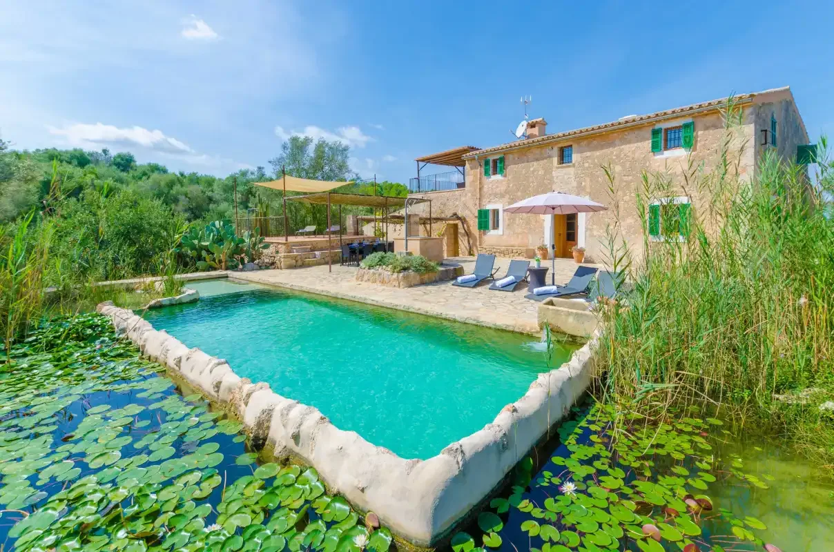 A stone house overlooks a swimming pool bordered by greenery and lily pads.