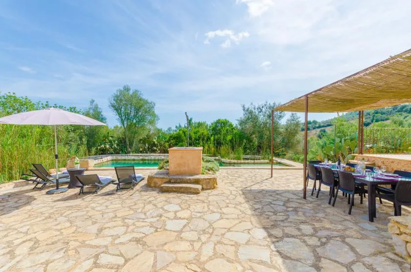 A stone patio surrounds a swimming pool, with outdoor seating beneath a shaded canopy.