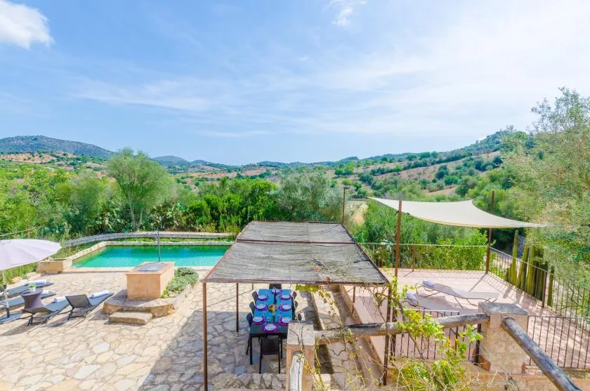 An outdoor patio with a shaded dining area overlooks a swimming pool and green hills.