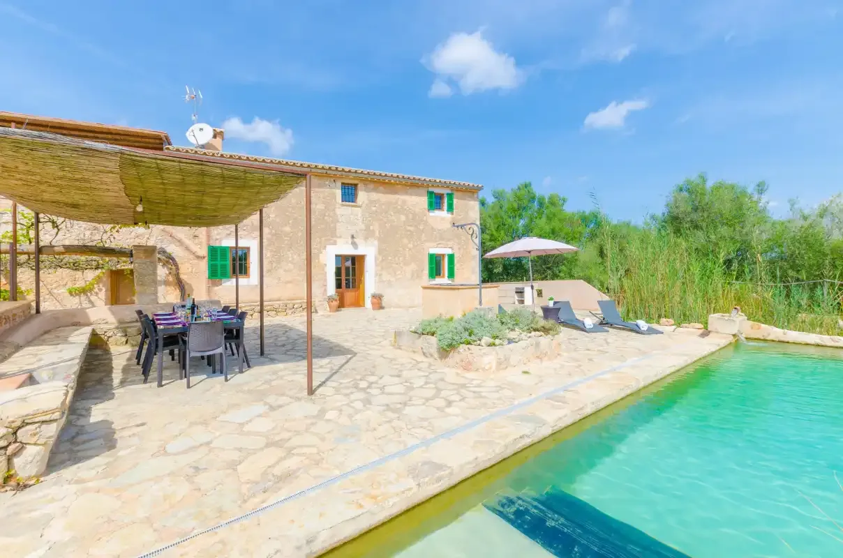A stone house and shaded patio overlook a swimming pool.