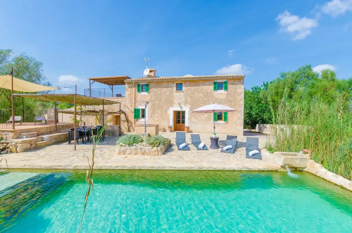 A stone house and patio overlook a swimming pool surrounded by greenery.