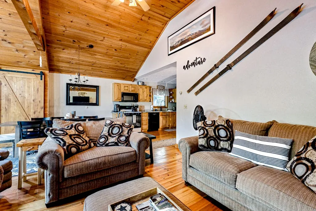 A living room with patterned cushions and a wooden ceiling, opening onto a kitchen.