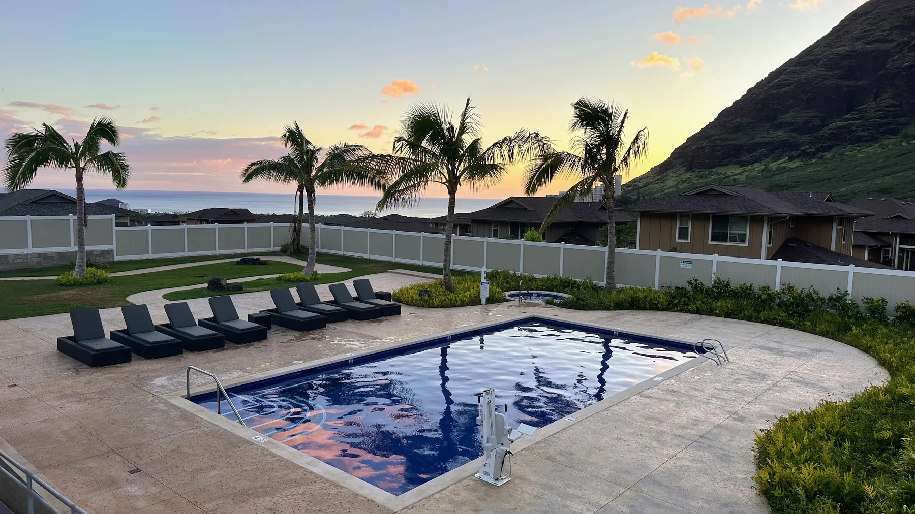 An outdoor swimming pool and lounge area sit beneath palm trees at sunset.
