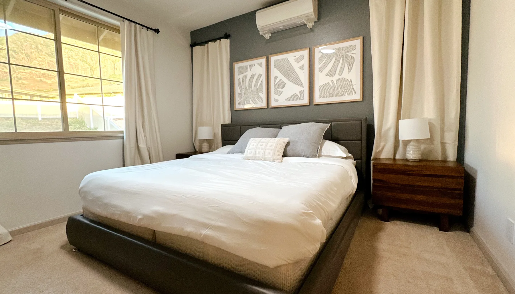Un dormitorio con una cama bien tendida, láminas enmarcadas y ventanas con cortinas.