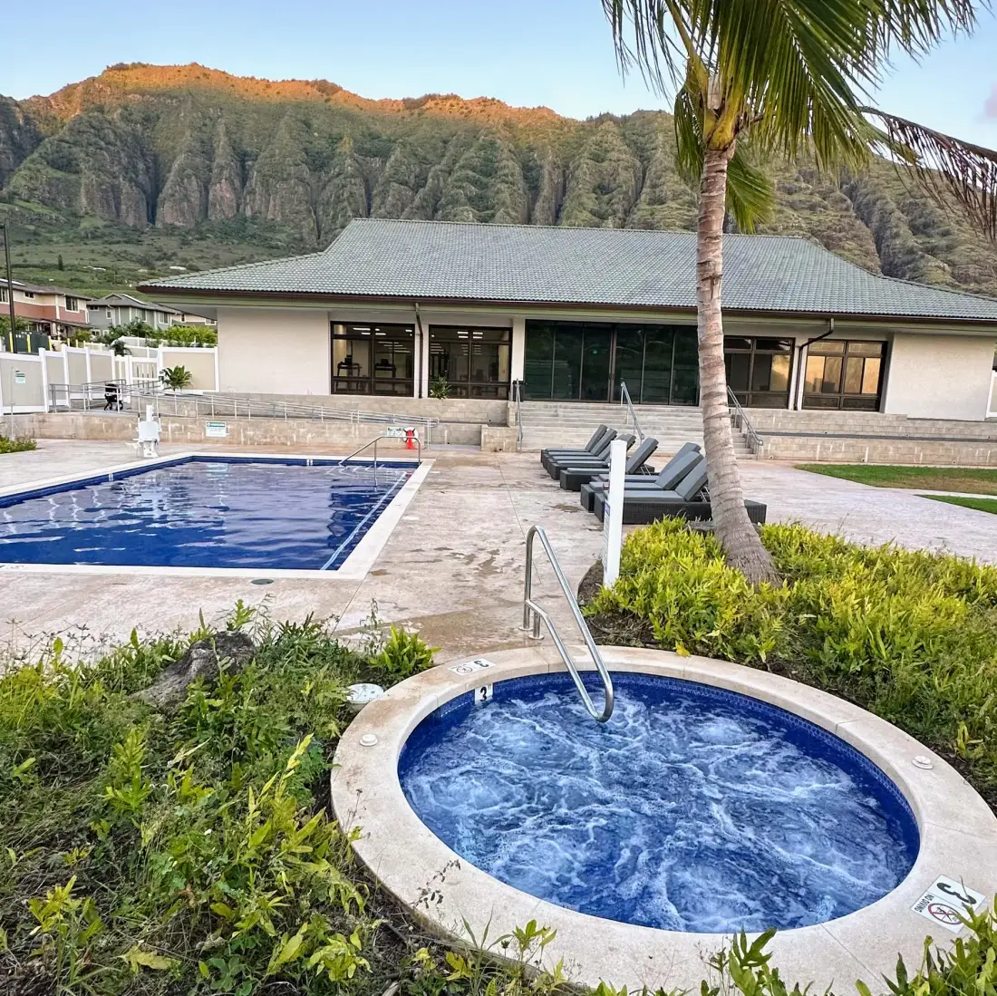 Una piscina al aire libre y un jacuzzi circular están junto a un edificio bajo, frente a una ladera boscosa.