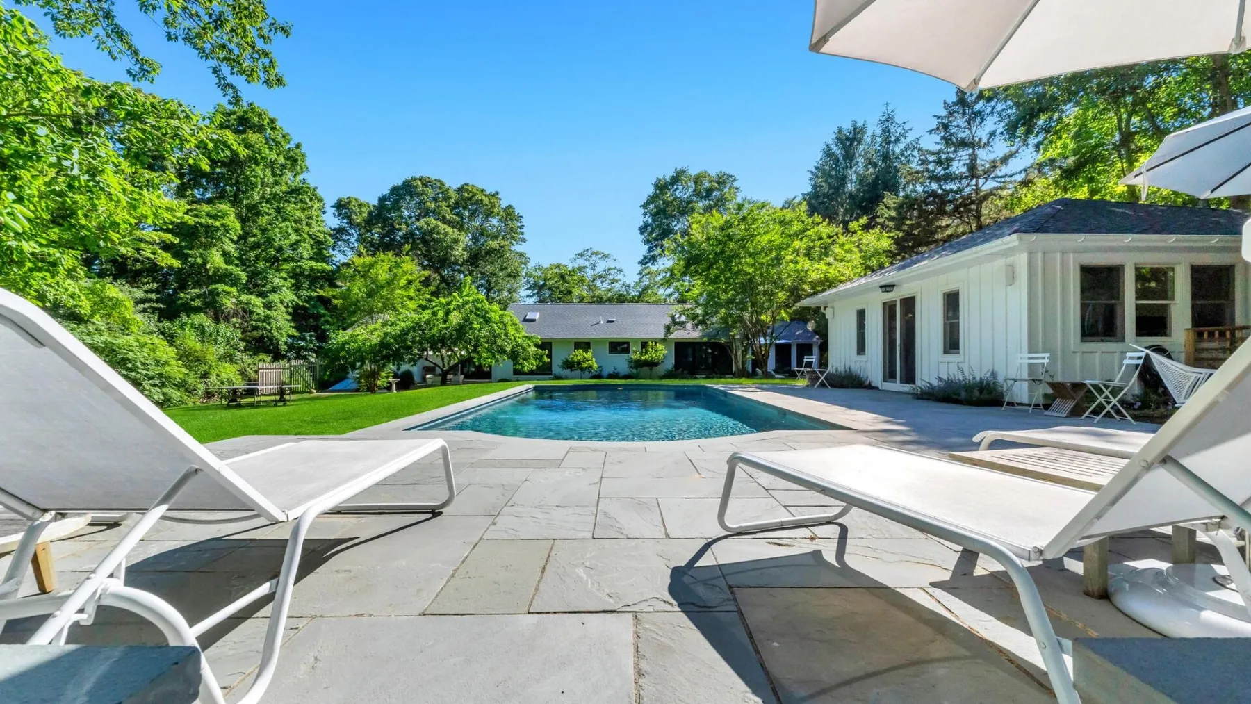 Una piscina y un patio pavimentado están junto a una casa blanca, rodeados de árboles.