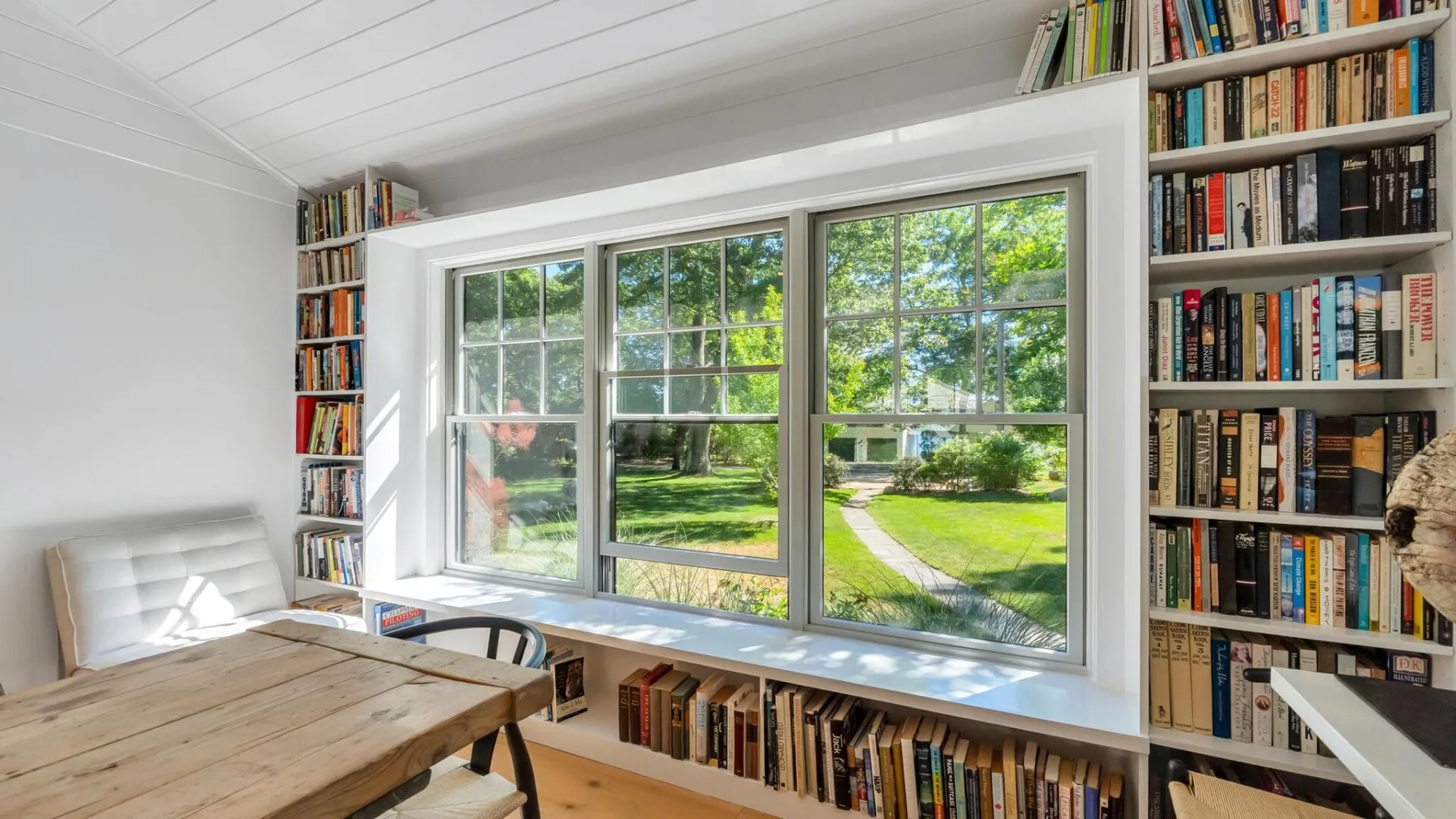 Un rincón de lectura luminoso con estanterías altas y amplias ventanas con vistas al jardín.