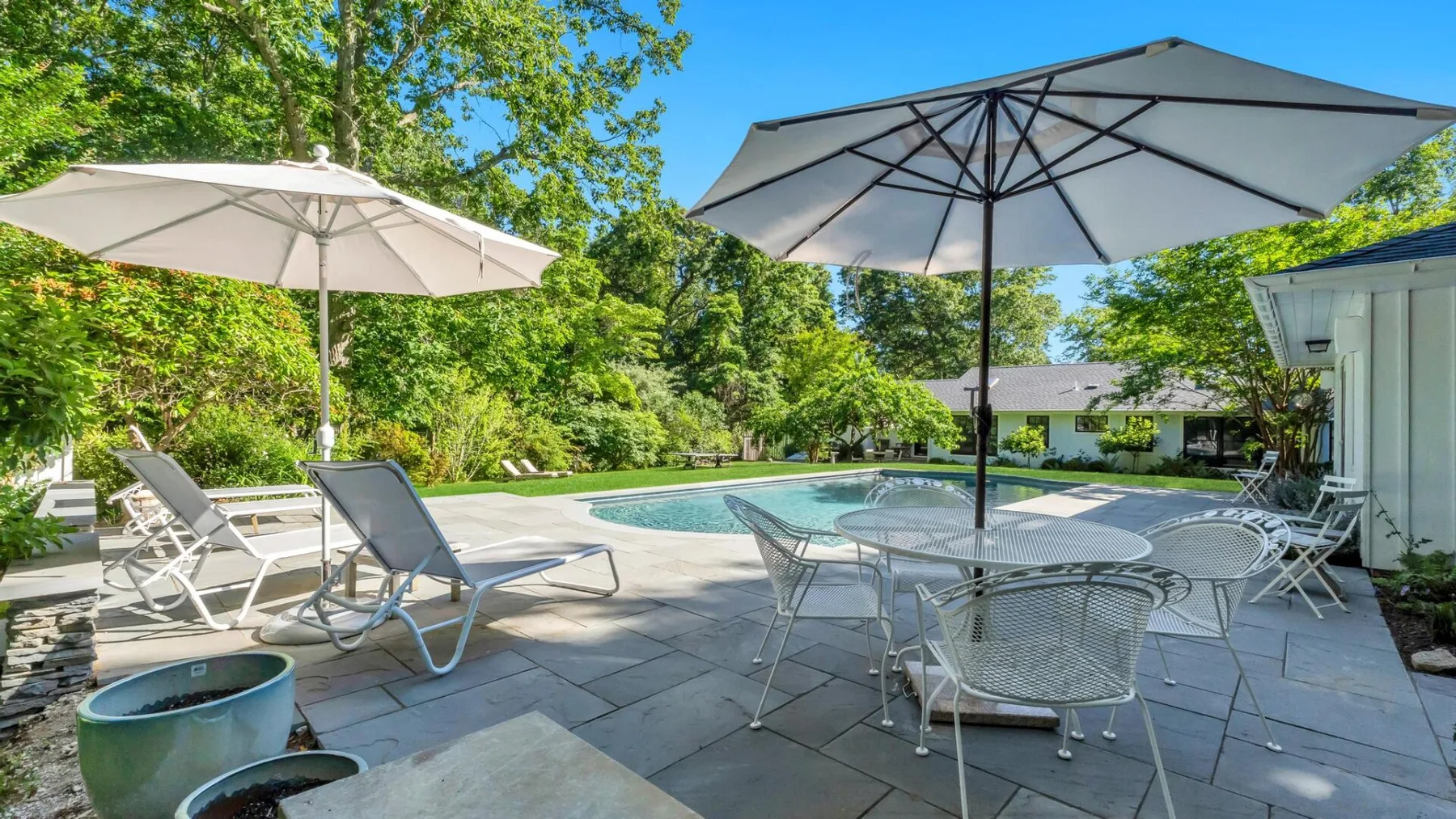 An outdoor patio with a swimming pool, shaded by umbrellas and surrounded by greenery.