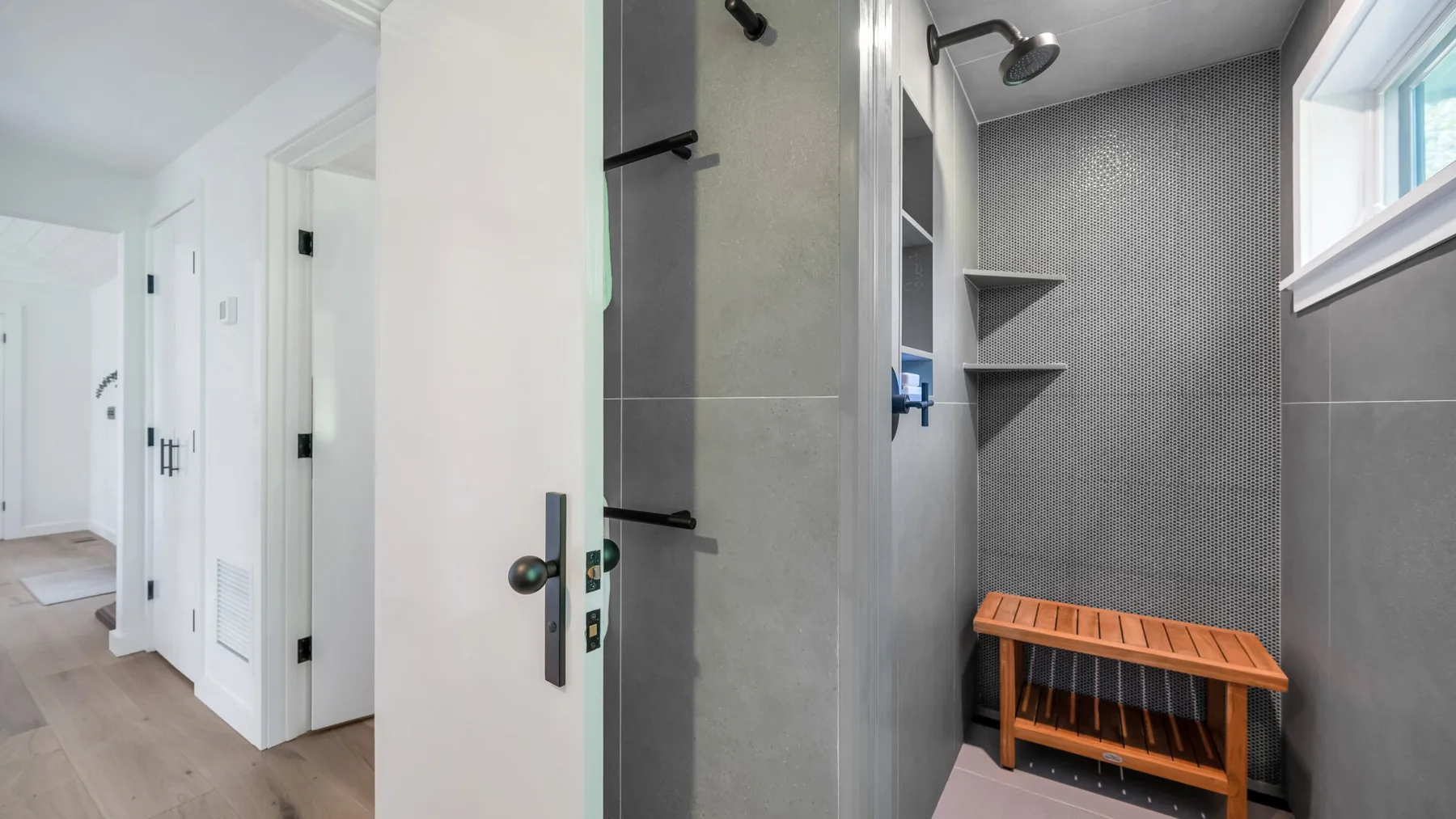 Una ducha con paredes de azulejos grises y un banco de madera junto a un pasillo blanco.