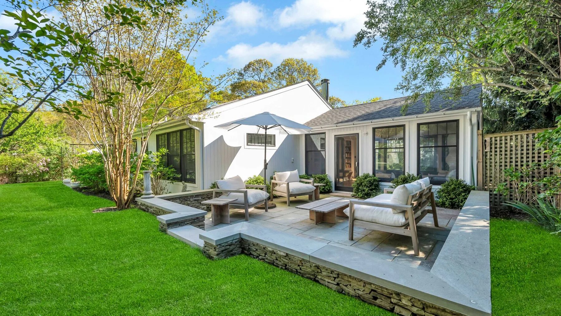 Un patio con asientos al aire libre junto a una casa blanca, rodeado de césped y árboles.