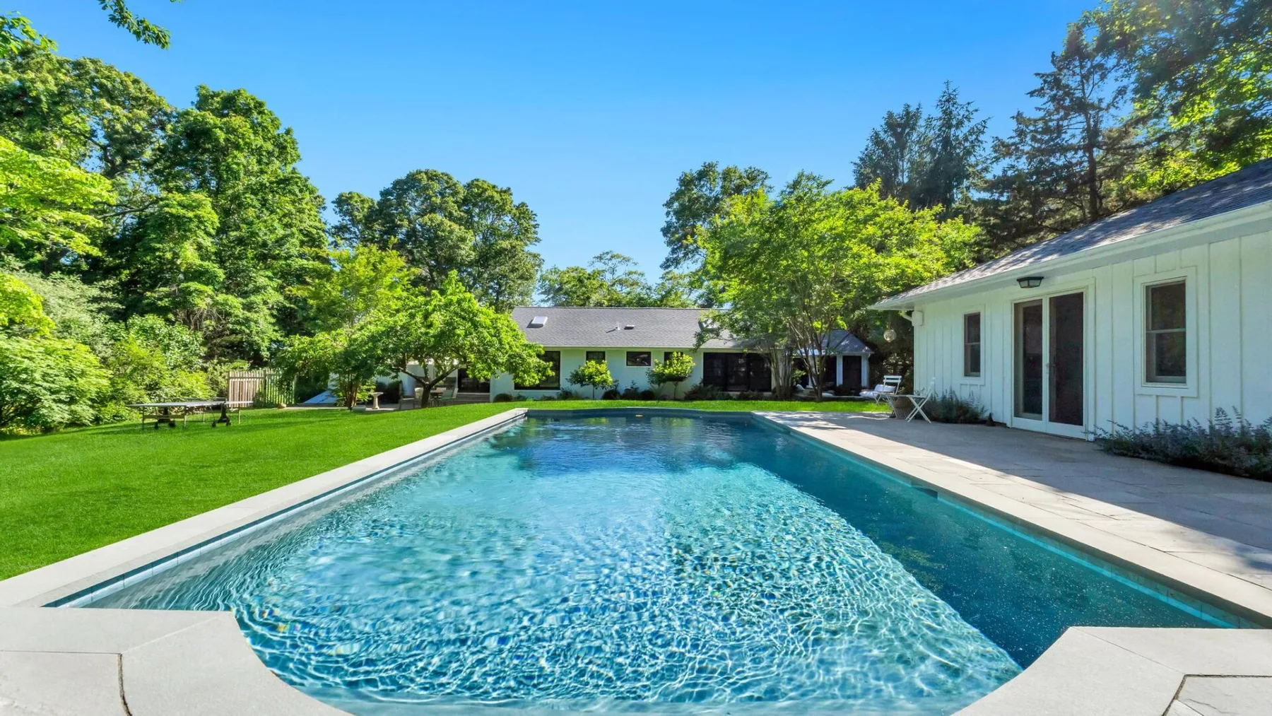 Outdoor swimming pool beside a white house, surrounded by green lawn and trees.