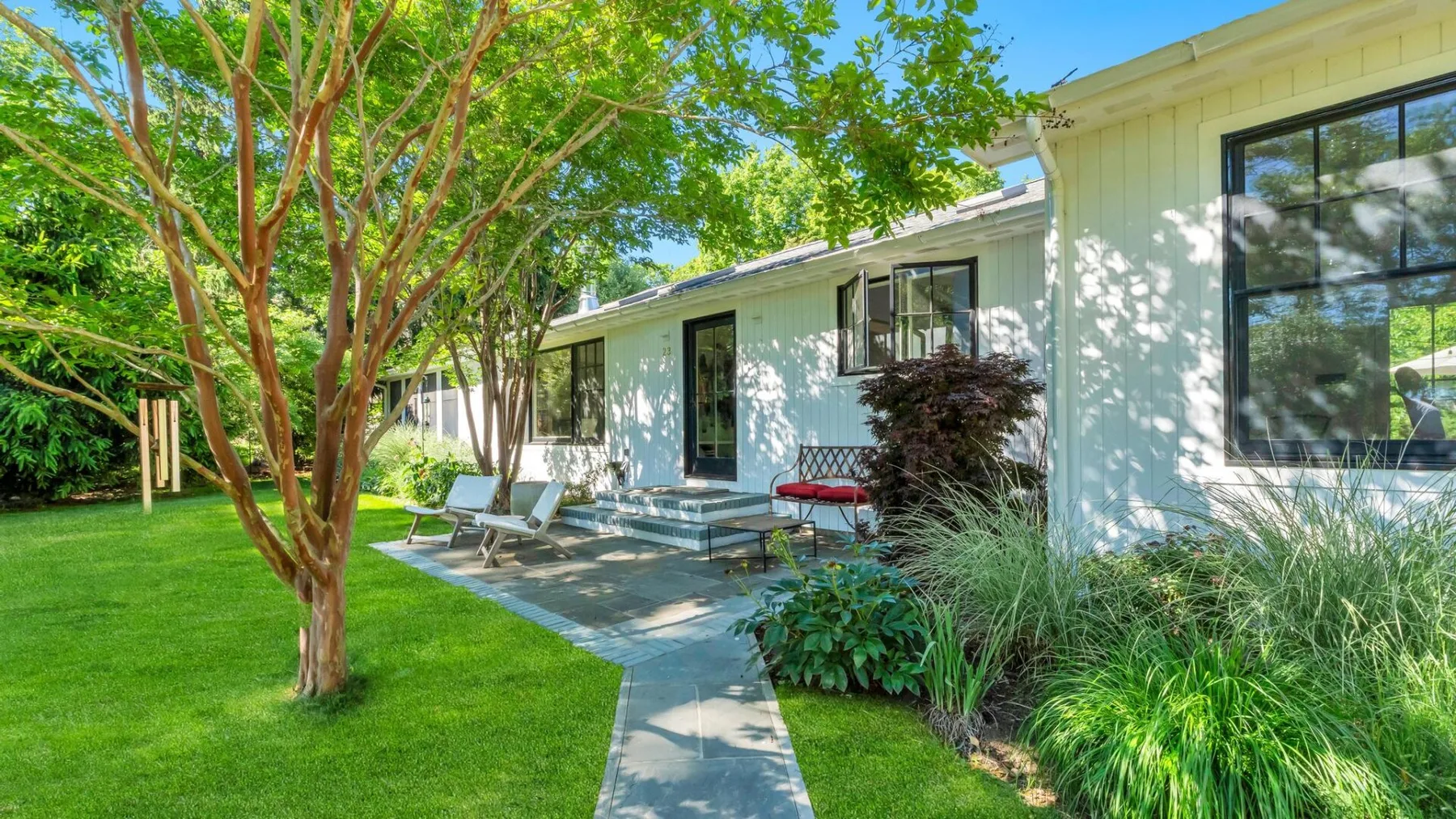 Un sendero de jardín conduce a una casa, con un árbol y plantas frondosas cerca.