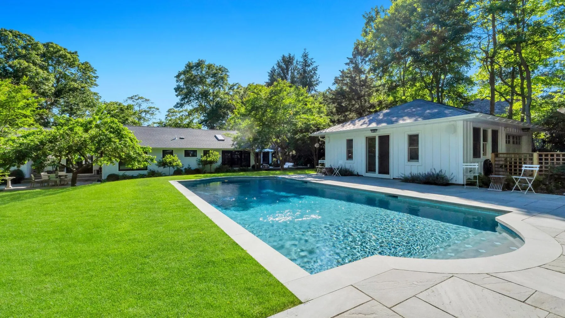 A swimming pool beside a white house, surrounded by green lawn and trees.