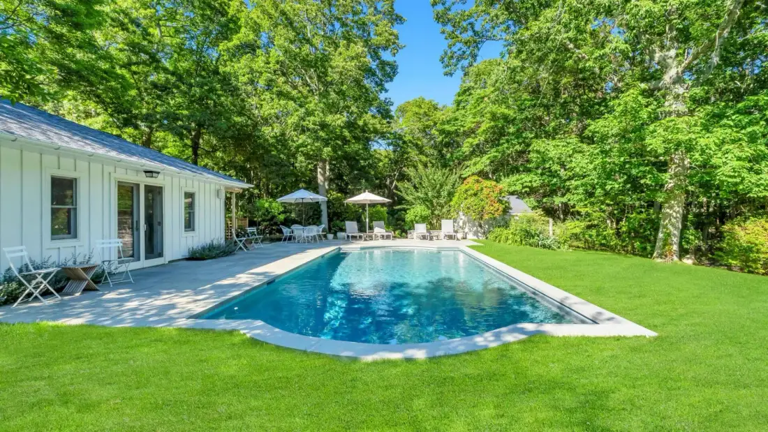 Una piscina al aire libre junto a una casa blanca, rodeada de césped y árboles.