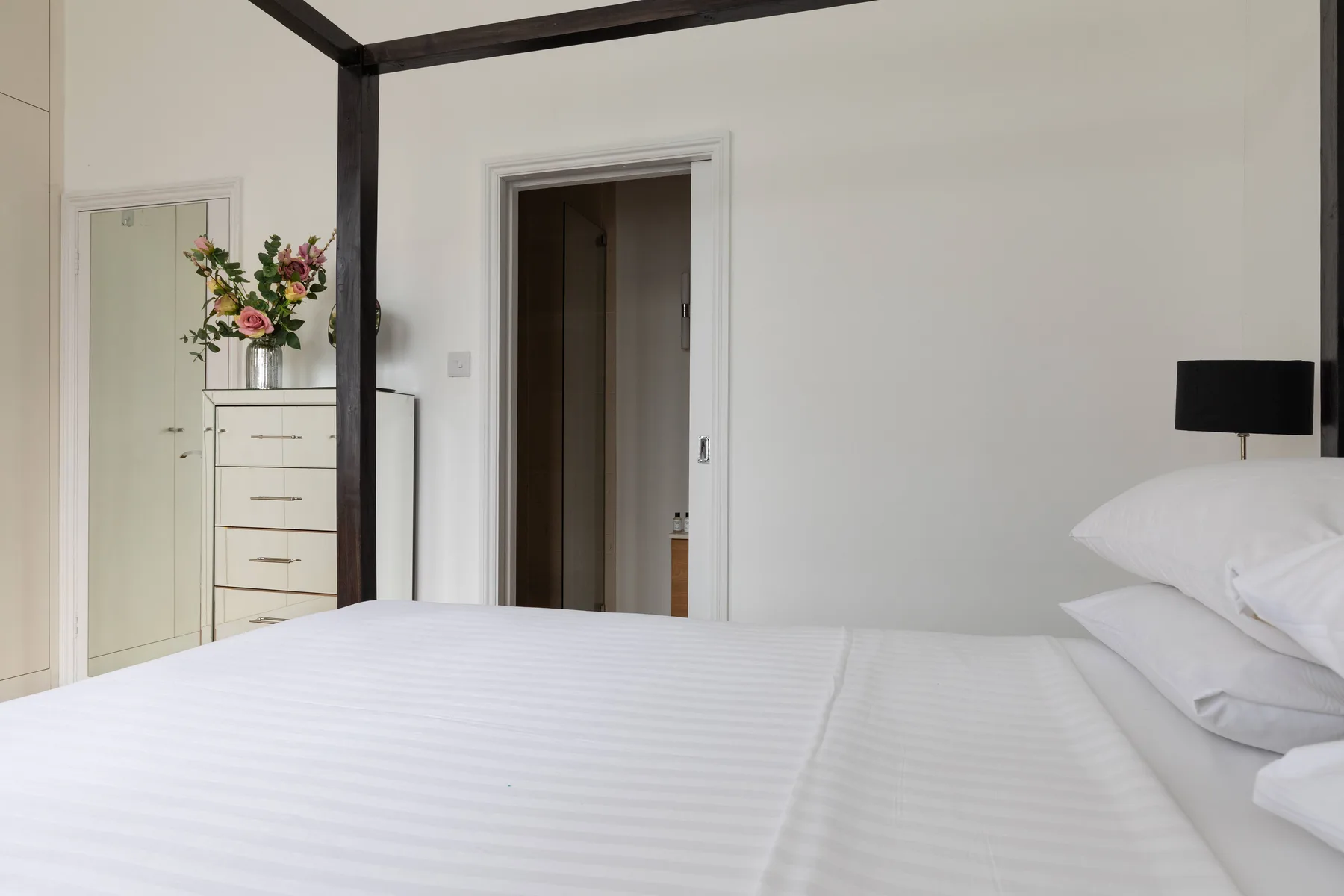 Un dormitorio luminoso con una cama bien tendida y flores sobre una cómoda.