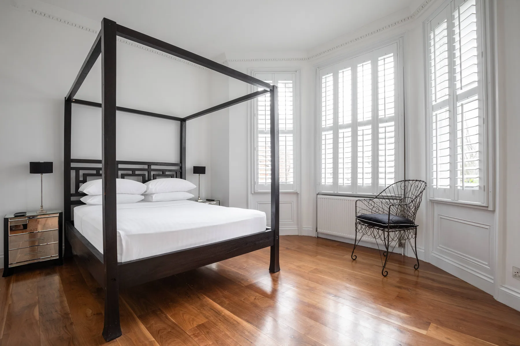 Un dormitorio luminoso con una cama con dosel oscura y ventanas altas con contraventanas.
