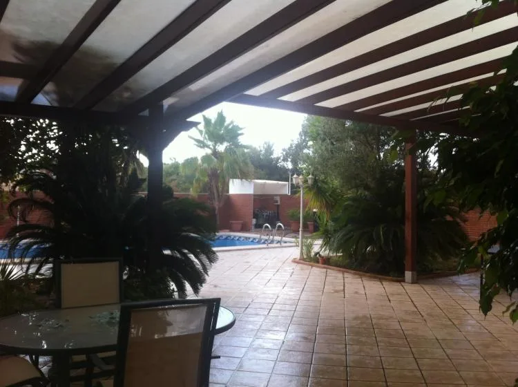 Un patio embaldosado bajo una pérgola cubierta da a una piscina y un jardín.