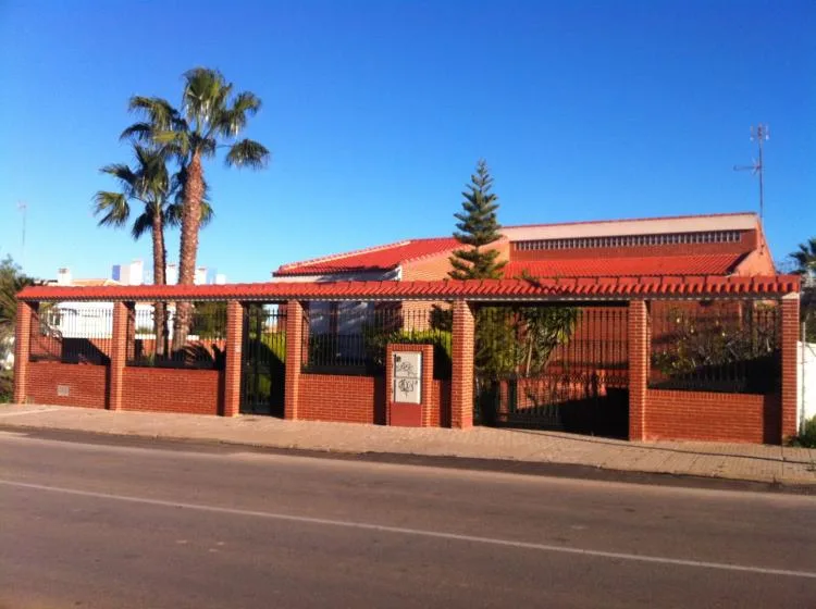 Una cerca baja de ladrillo y metal está frente a un edificio de techo rojo bajo un cielo despejado.