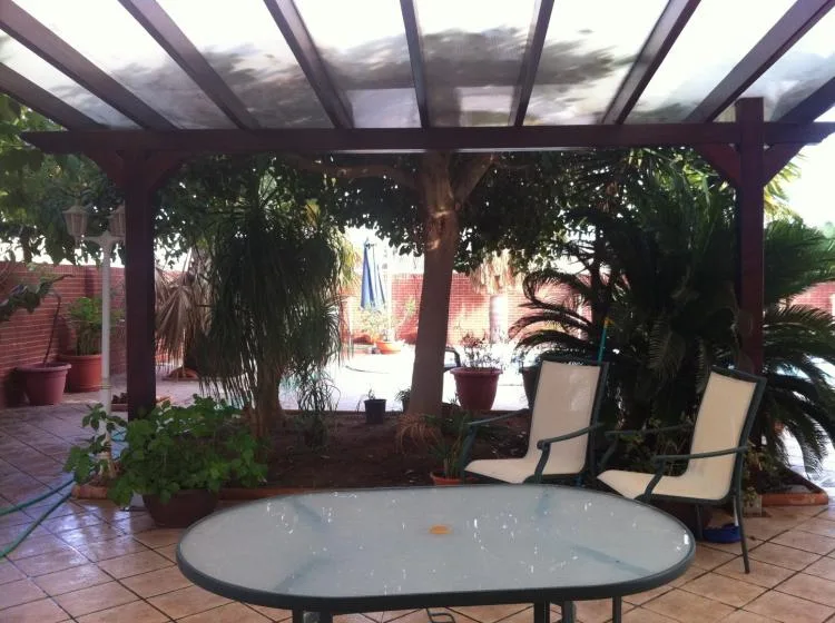 Un patio embaldosado tiene una mesa de vidrio, asientos al aire libre y plantas frondosas bajo una pérgola.