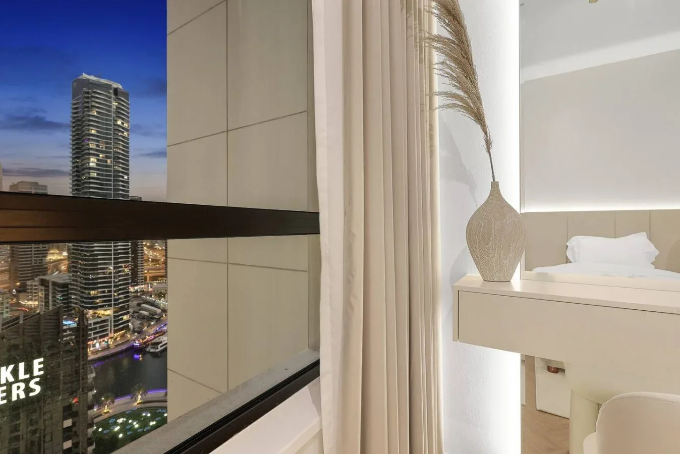 A bright bedroom with a bed beside a large window overlooking city buildings.
