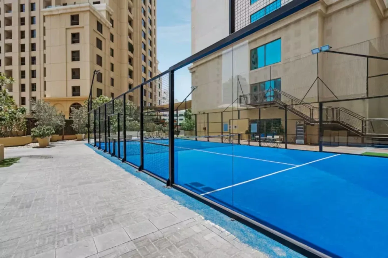 An outdoor blue court is enclosed by a tall black fence beside buildings.