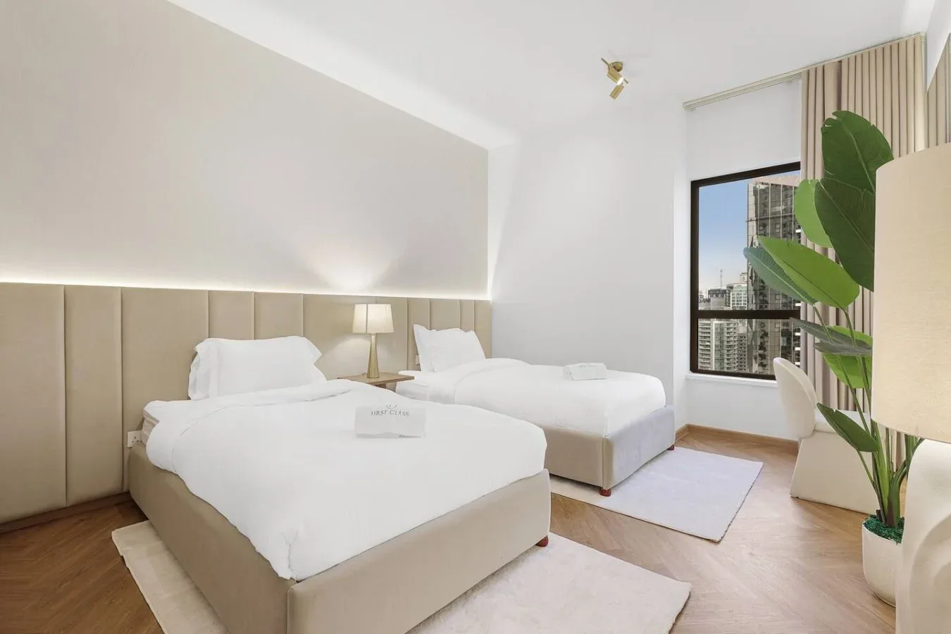 A bright bedroom with two beds, a bedside lamp, and a large window.