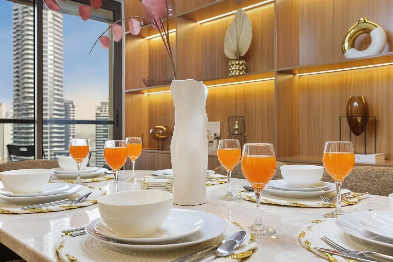 A dining table is set with white dishes and orange drinks beside a window.