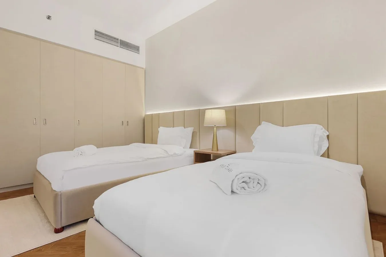 A bedroom with two beds, neutral walls, and a bedside lamp.