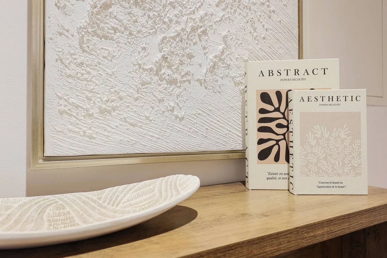 A framed textured artwork hangs above a wooden console with decorative books and a shallow dish.