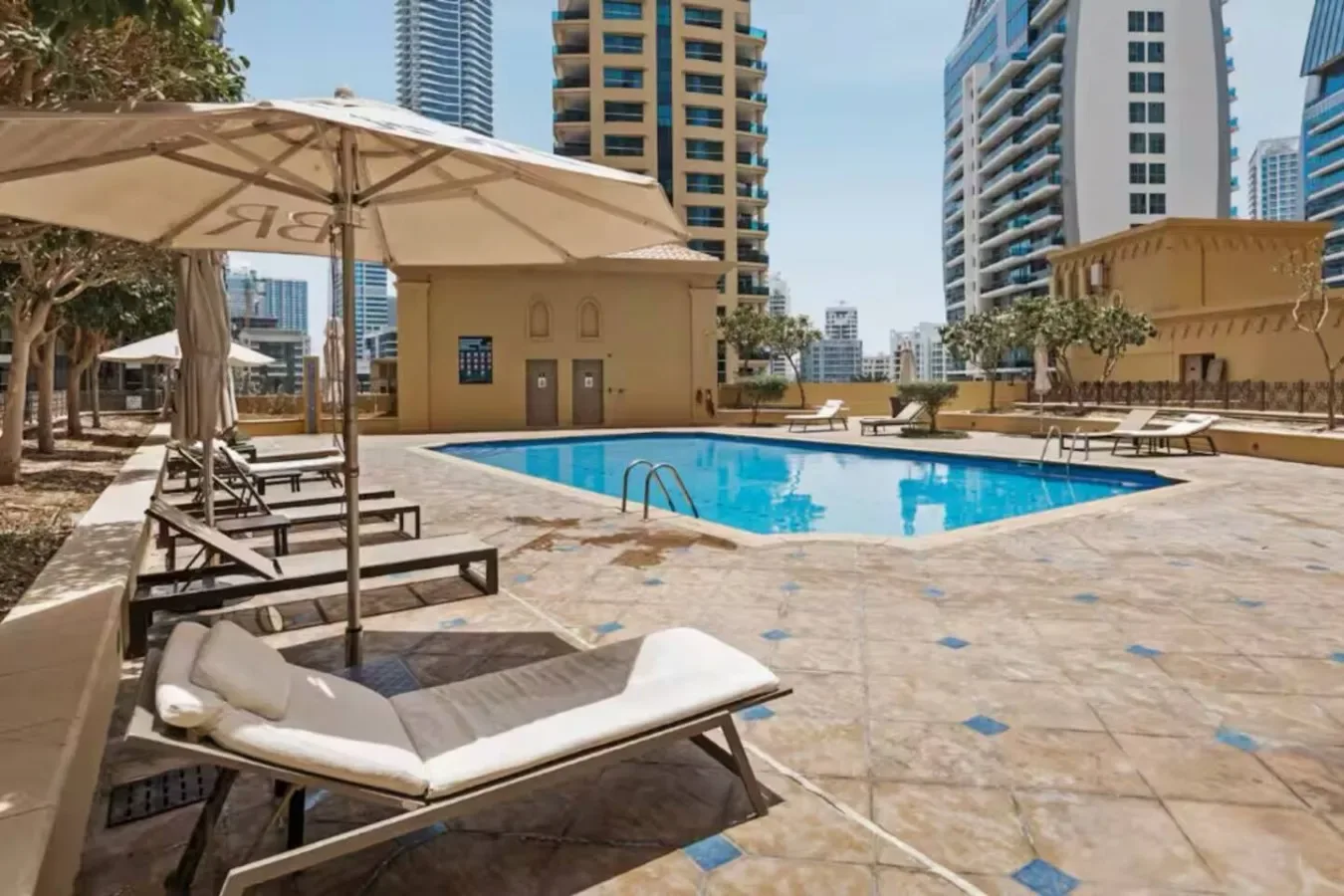 An outdoor swimming pool sits beside shaded loungers and tall apartment buildings.