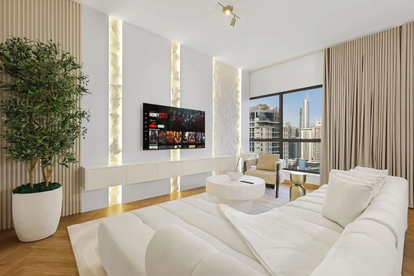 A bright living room features white seating, a wall-mounted television, and large windows.
