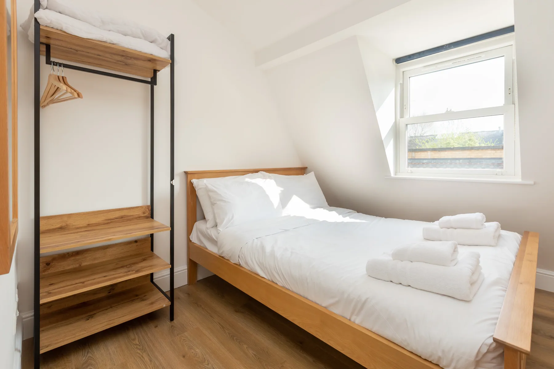 A bright bedroom with a wooden bed and open clothes rack beside a window.