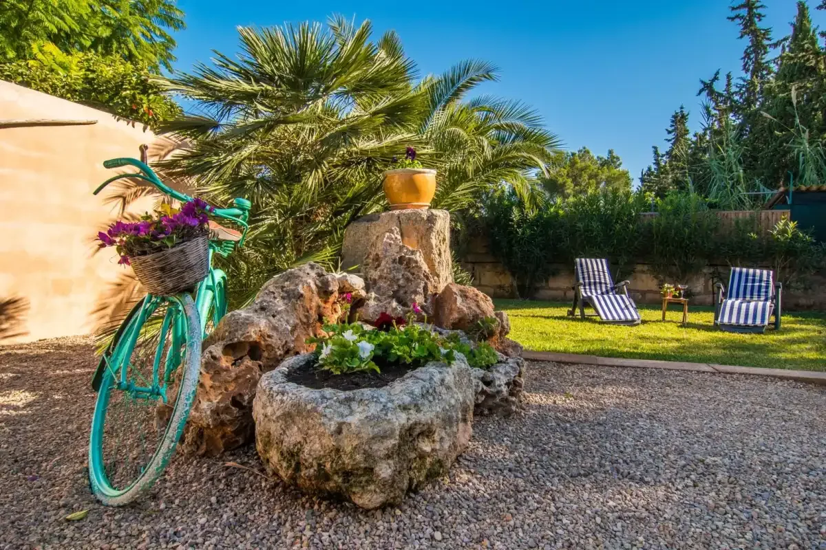 Un jardín soleado con una fuente de piedra, una bicicleta turquesa y tumbonas a rayas.