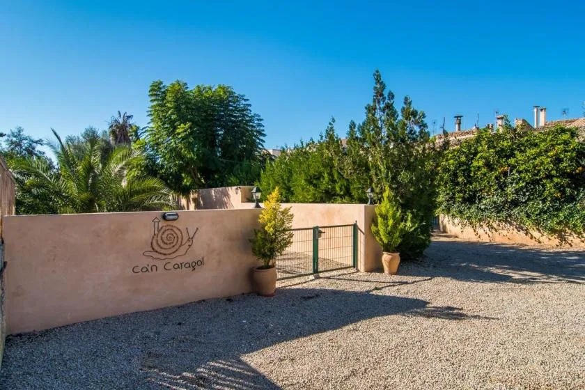 Una entrada de patio de grava bordeada por un muro bajo, una puerta y árboles frondosos.