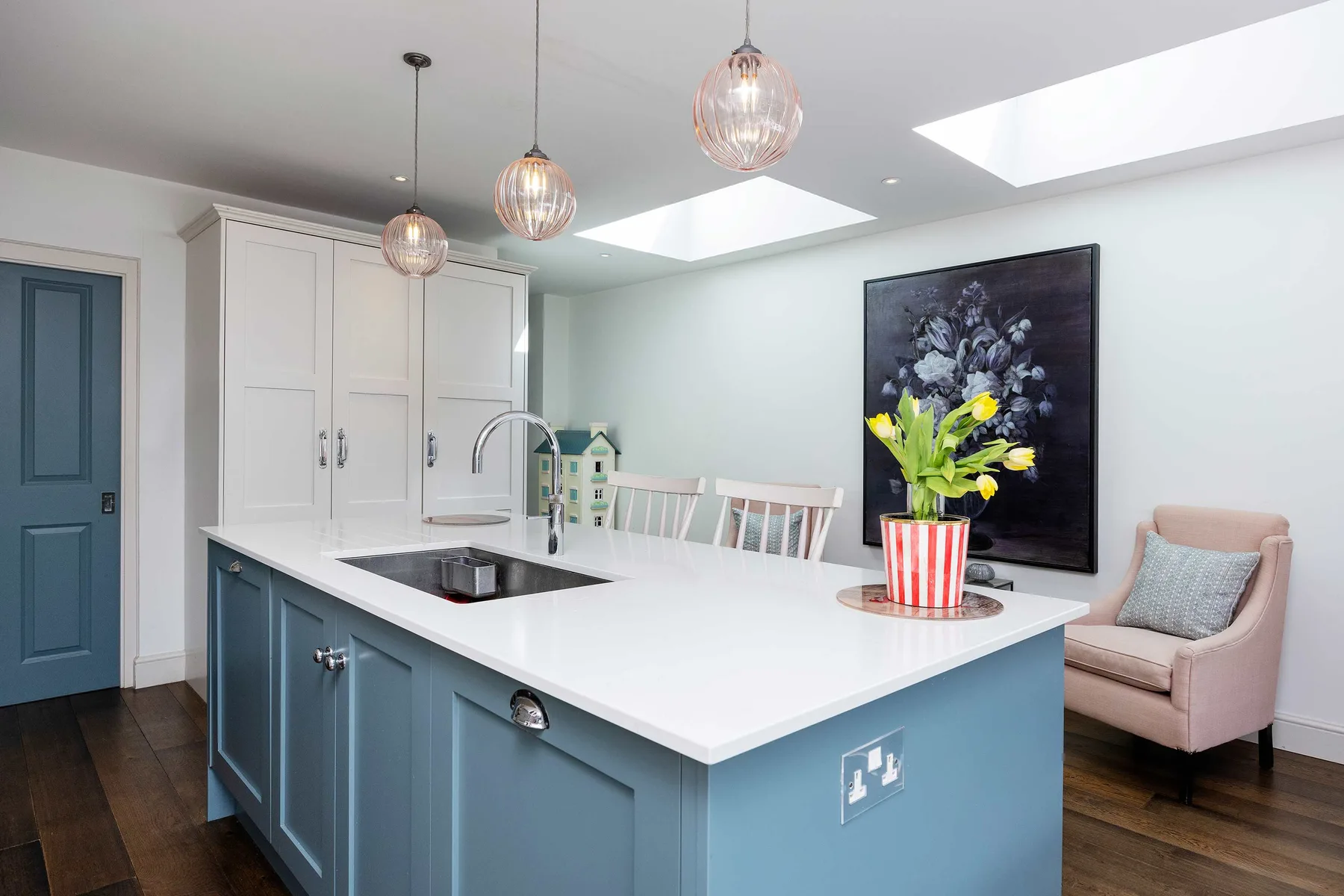 A bright kitchen with a large blue island, pendant lights, and a pink armchair.