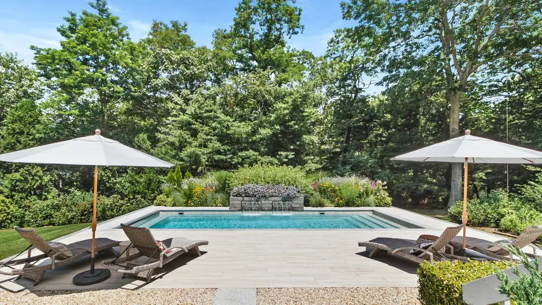 An outdoor swimming pool sits beside a wooden deck with lounge seating and umbrellas.