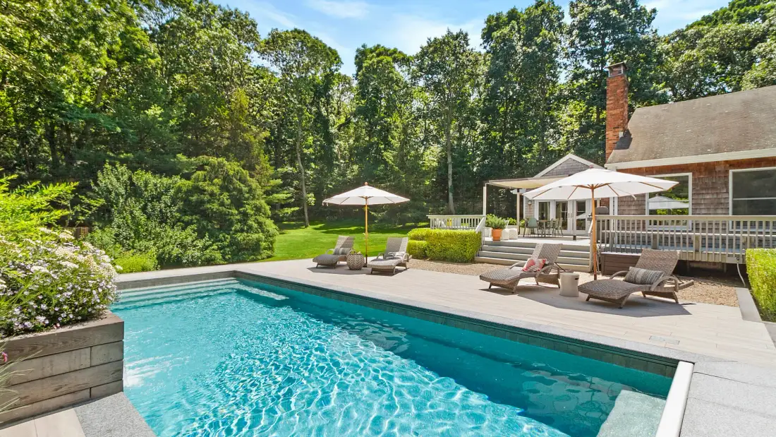 An outdoor swimming pool beside a deck with lounge seating and a wooded backdrop.