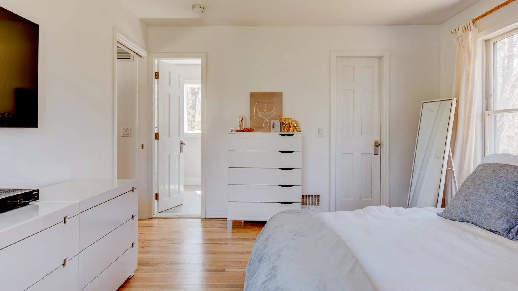 Un dormitorio luminoso con una cama, una cómoda blanca y ventanas grandes.