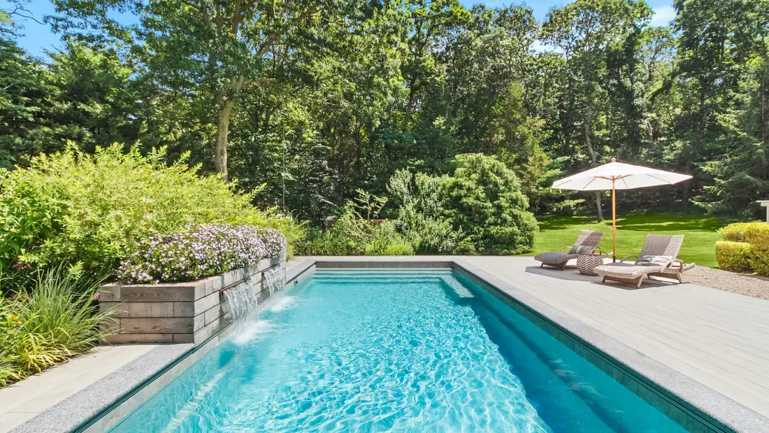 An outdoor swimming pool is surrounded by a wooden deck, lounge seating, and greenery.