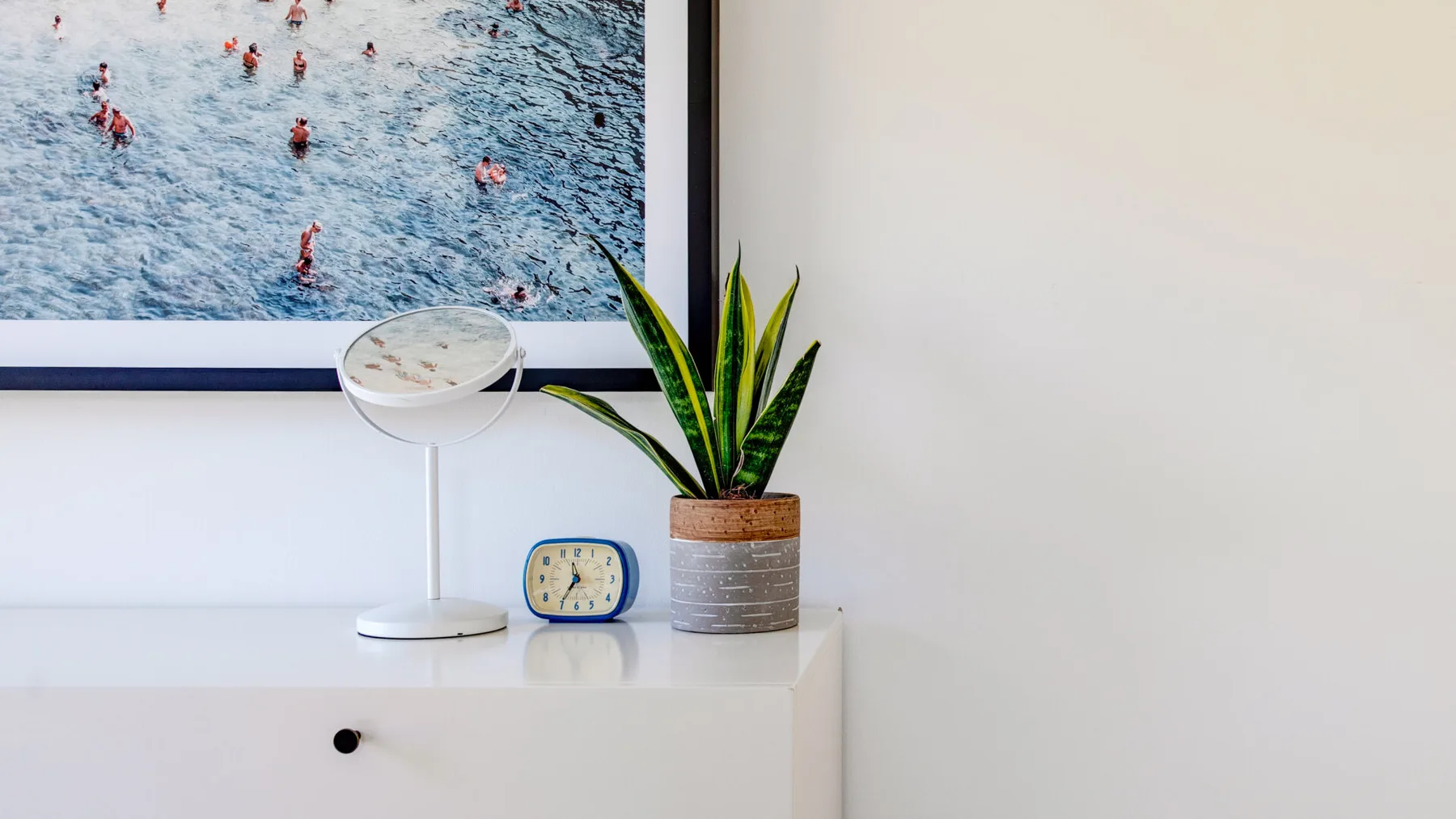 Una cómoda de dormitorio muestra una planta en maceta, un reloj y una lámpara bajo una obra enmarcada.