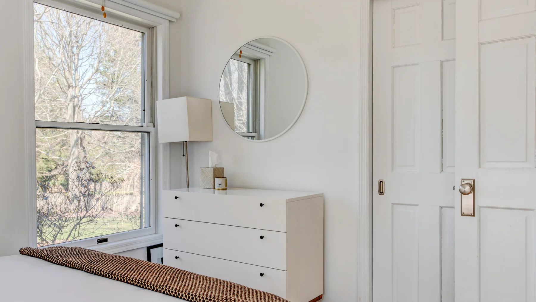Un dormitorio luminoso con una cómoda y un espejo redondo junto a una ventana grande.