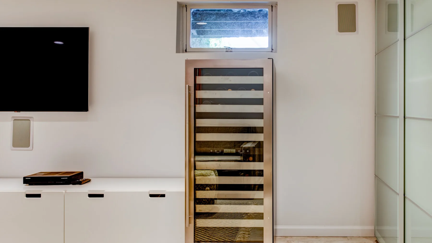Un interior luminoso con un televisor en la pared, un mueble bajo y una ventana estrecha.