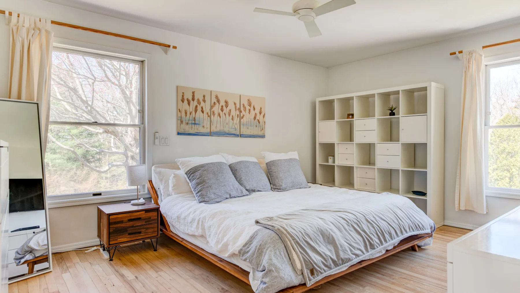 Un dormitorio luminoso con una cama, ventanas grandes y una estantería blanca con compartimentos.