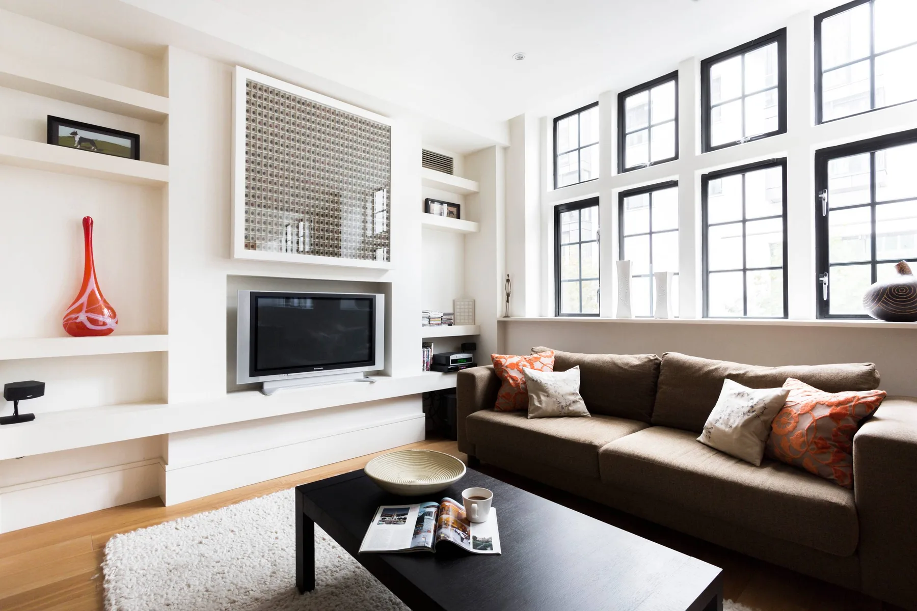 Una sala luminosa con un sofá seccional oscuro bajo grandes ventanas de cuadrícula.