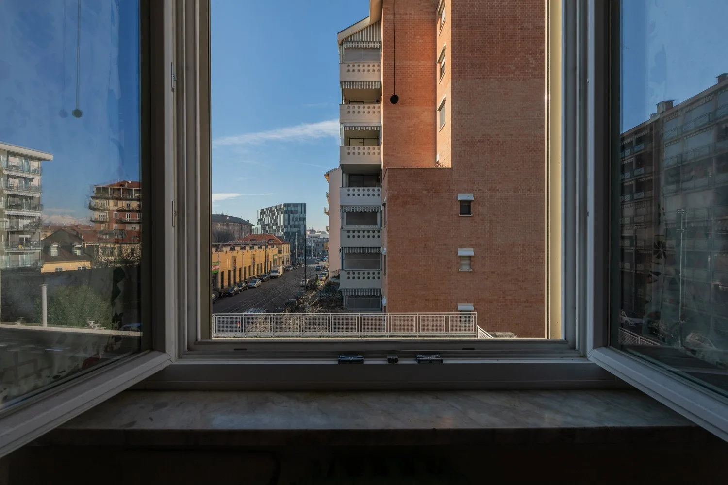 Una ventana abierta enmarca una vista de edificios de apartamentos y una calle urbana.