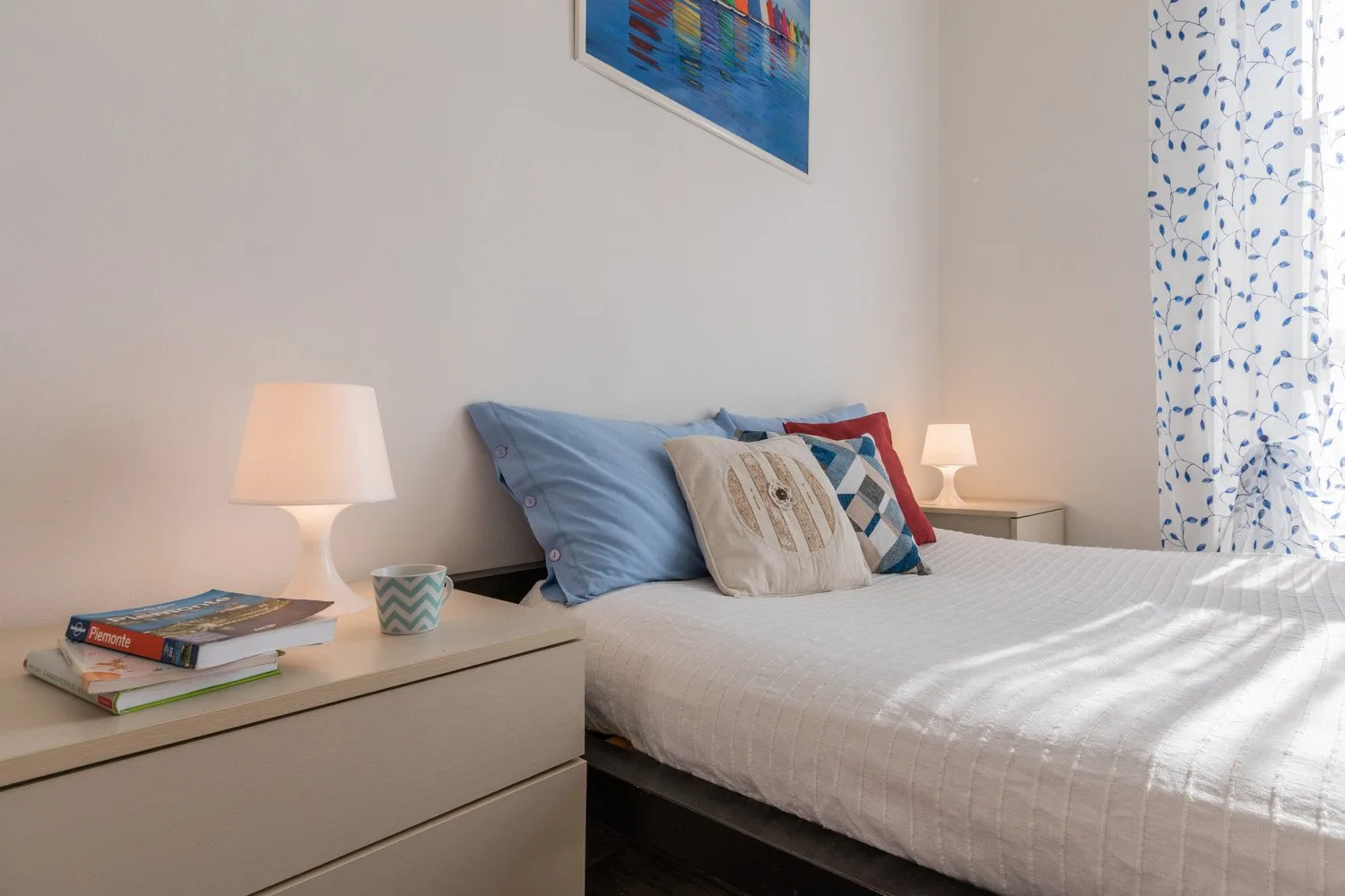 Un dormitorio con una cama tendida, lámparas de noche y cortinas estampadas.