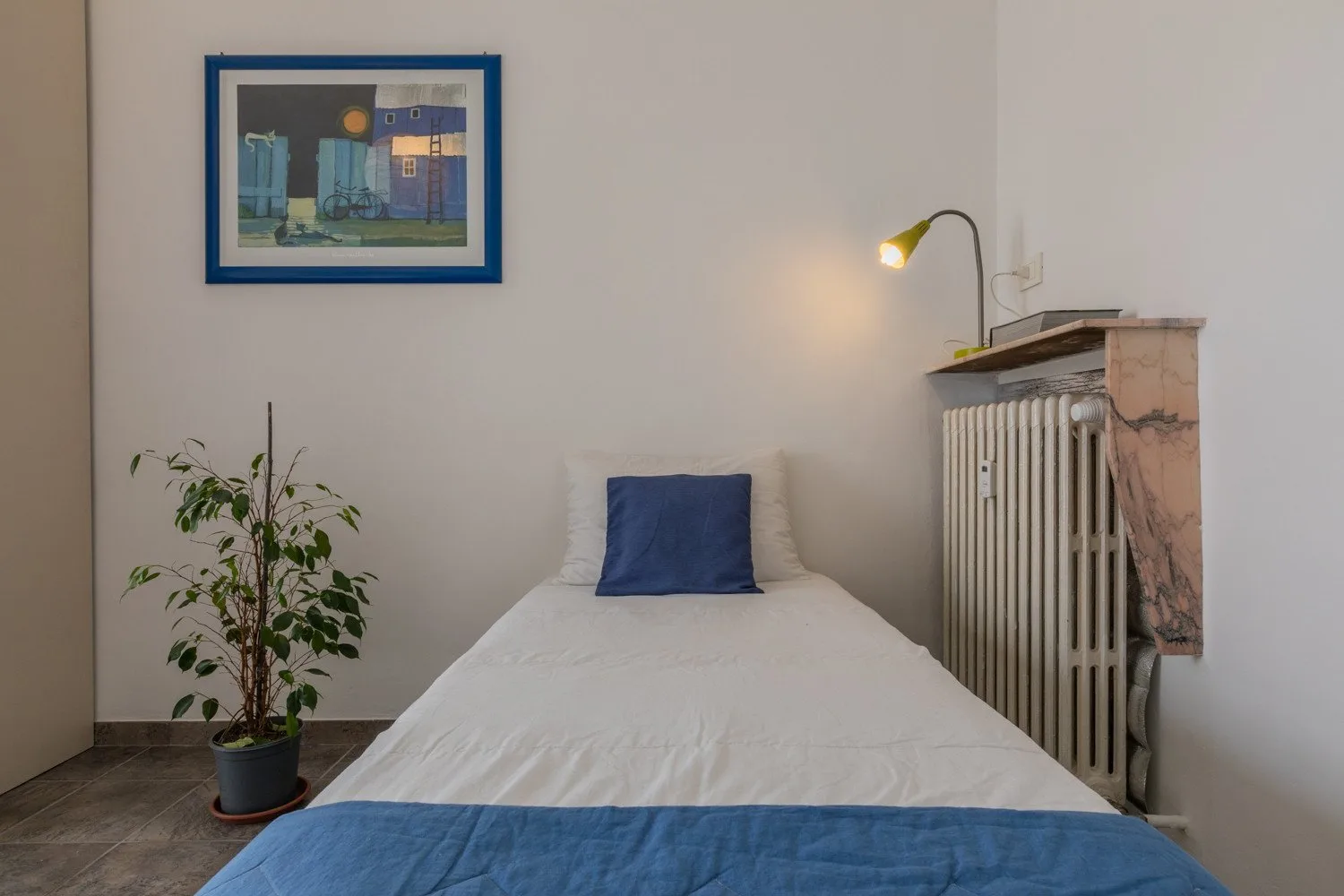 Una cama individual con ropa de cama blanca y una manta azul queda bajo un cuadro enmarcado.